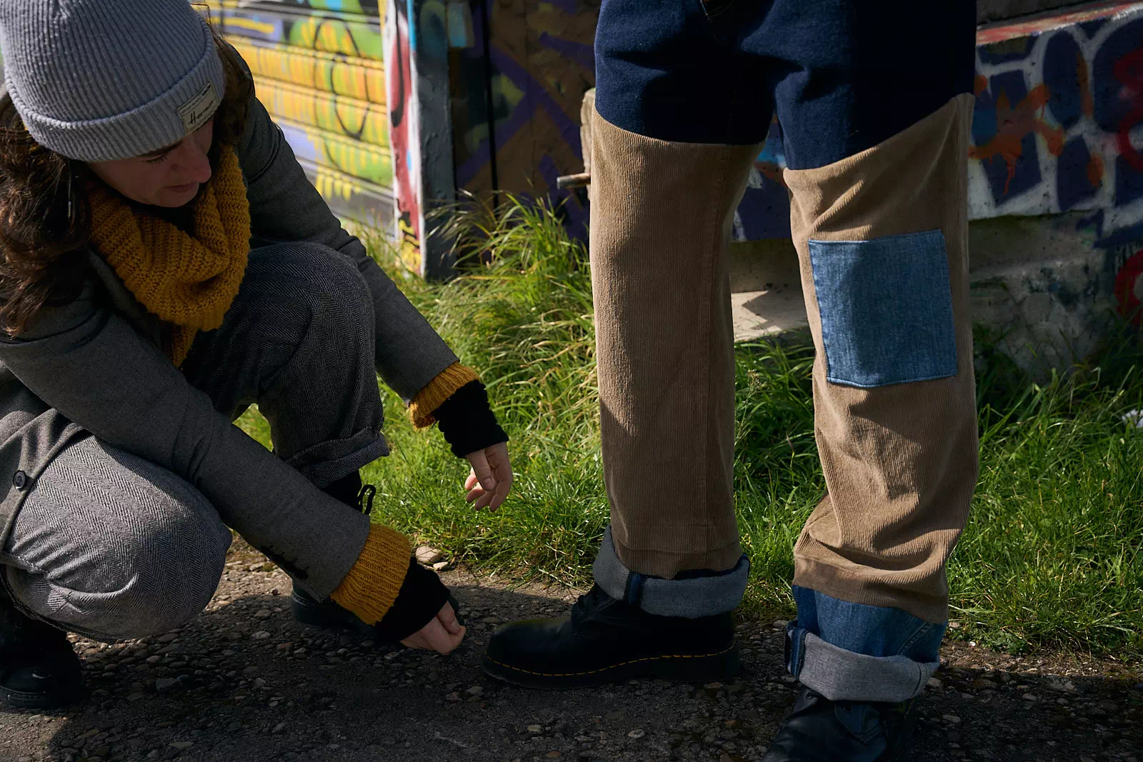Lucie de Turcou est accroupie sur la gauche de l'image et elle réajuste la tenue du mannequin sur la droite. Il porte un pantalon upcyclé mixant jeans et velours. En fond il y a un bâtiment abandonné et tagué